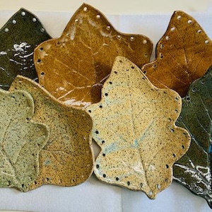 May include: A set of seven ceramic leaf-shaped dishes in various shades of green, brown, and beige. The dishes have a textured surface and are decorated with small holes around the edges.
