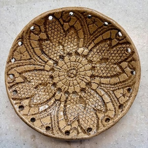 May include: A handmade, round ceramic plate with a detailed, lace-like pattern in shades of brown. The plate features a series of evenly spaced holes around the rim. The design is reminiscent of vintage doilies.
