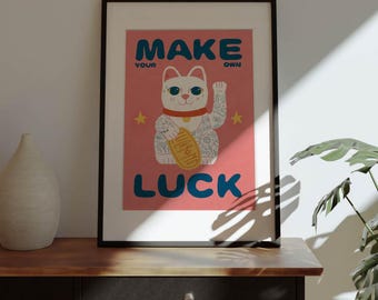 May include: A pink poster with a white cat with tattoos and a gold coin. The cat is sitting with its paw raised. The text on the poster reads "MAKE YOUR OWN LUCK".