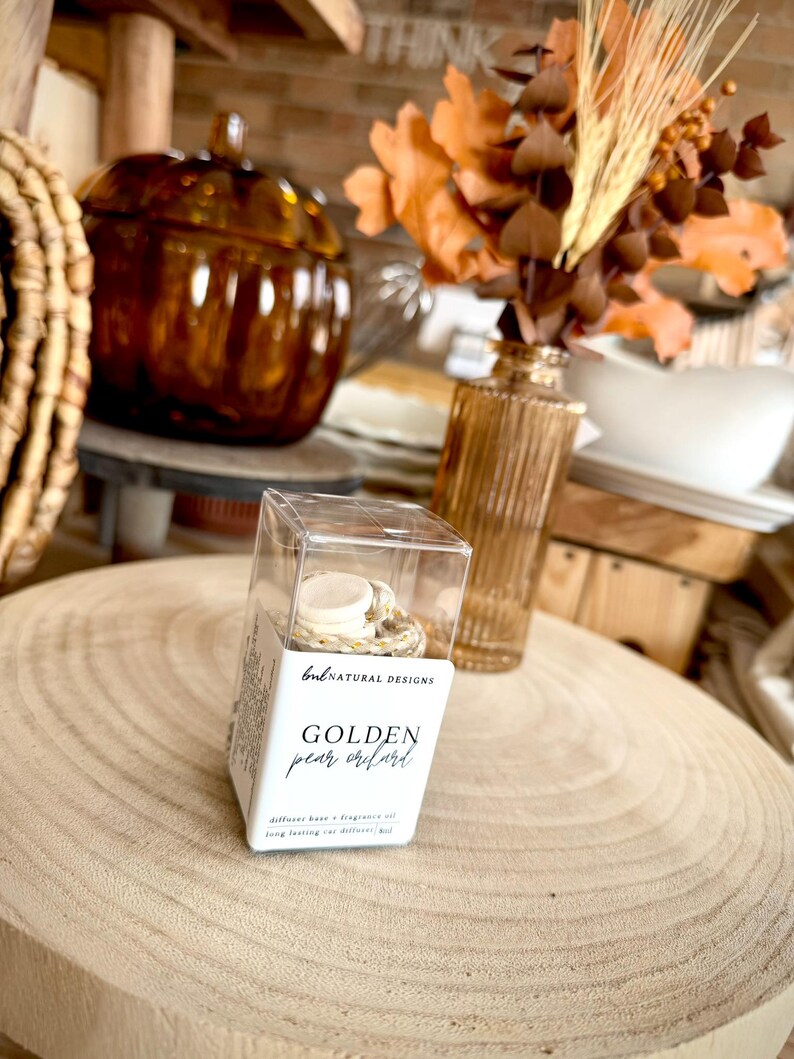 May include: A clear plastic box containing a car diffuser with a white cap and a floral patterned base. The label reads BNL NATURAL DESIGNS, GOLDEN pear orchard, and diffuser base & fragrance oil, long lasting car diffuser 8ml.
