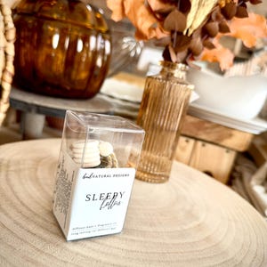 May include: A clear plastic box containing a car diffuser with white wooden beads and a label that reads BNL NATURAL DESIGNS SLEEPY HOLLOW diffuser base and fragrance oil long lasting car diffuser 8ml, positioned on a light wood surface.