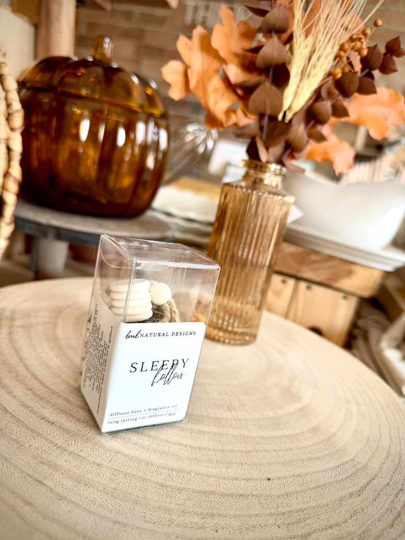 May include: A clear plastic box containing a car diffuser with white wooden beads and a label that reads BNL NATURAL DESIGNS SLEEPY HOLLOW diffuser base and fragrance oil long lasting car diffuser 8ml, positioned on a light wood surface.