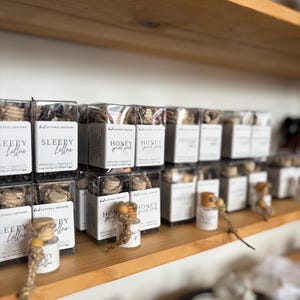 May include: A collection of small, clear glass car diffusers with wooden caps and hanging cords, packaged in square boxes labeled with brand names like Sleepy Hollow and Honey Spiced Pear, arranged on a wooden shelf in a retail display.