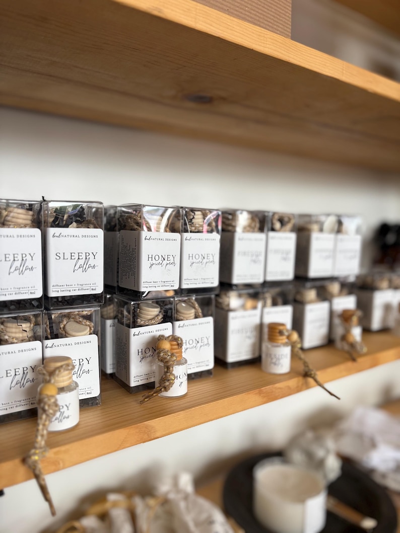 May include: A collection of small, clear glass car diffusers with wooden caps and hanging cords, packaged in square boxes labeled with brand names like Sleepy Hollow and Honey Spiced Pear, arranged on a wooden shelf in a retail display.