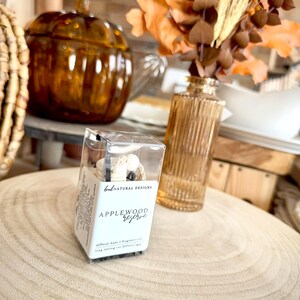 May include: A clear rectangular box containing a car diffuser with white wooden beads and black cord. The label reads BNL NATURAL DESIGNS and APPLEWOOD reserve. The product is positioned on a light wood surface with a glass vase and pumpkin.