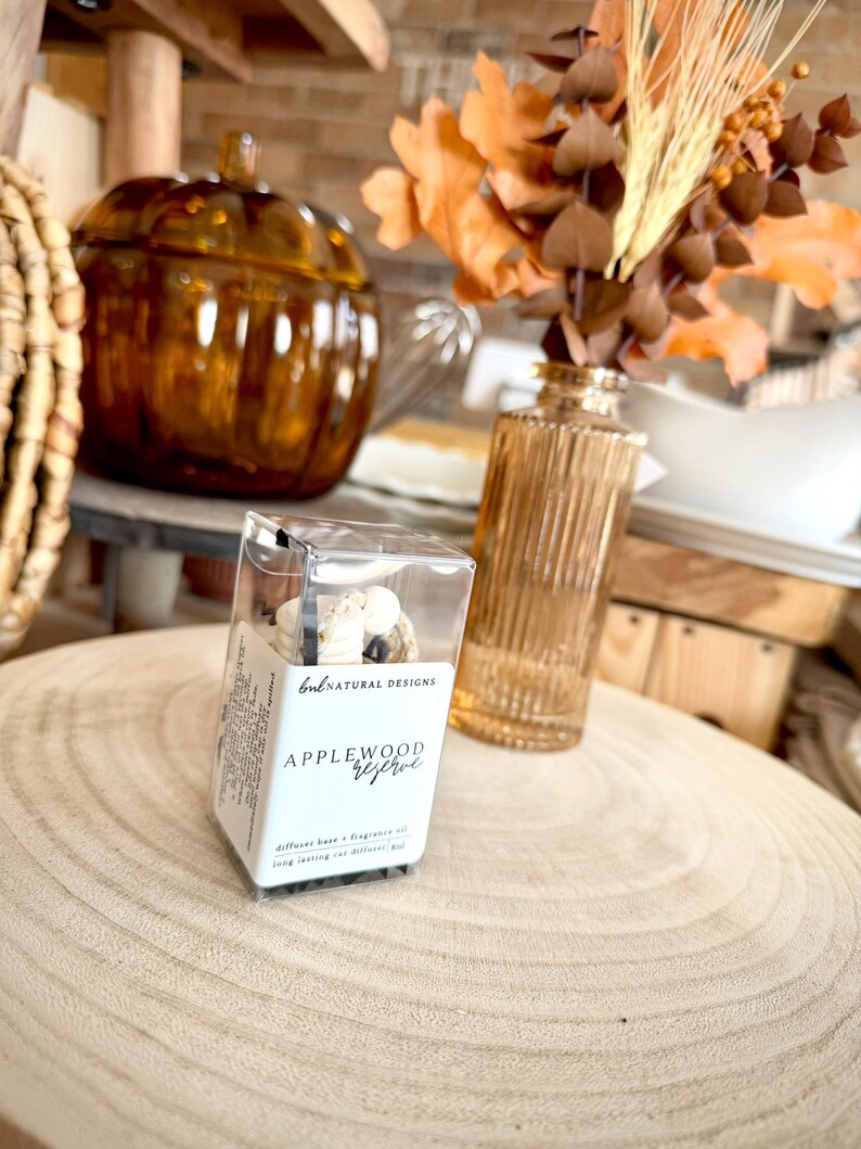May include: A clear rectangular box containing a car diffuser with white wooden beads and black cord. The label reads BNL NATURAL DESIGNS and APPLEWOOD reserve. The product is positioned on a light wood surface with a glass vase and pumpkin.