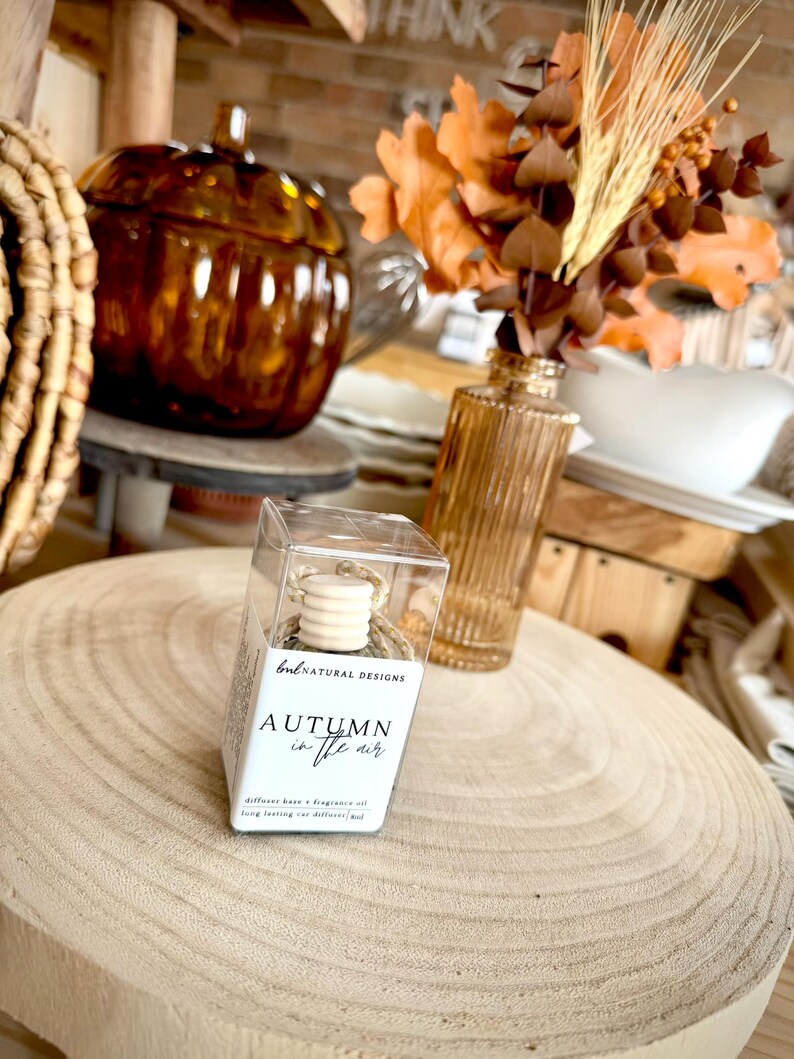 May include: A clear plastic box containing a car diffuser with a wooden cap and twine. The white label features the text "bnl NATURAL DESIGNS" and "AUTUMN in the air" with "diffuser base + fragrance oil" and "long lasting car diffuser 8ml".