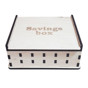 May include: A light-colored wooden savings box with the words "Savings box" engraved on the lid. The box has a rectangular shape with black trim and small rectangular cutouts along the sides.