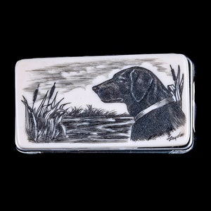 Puede incluir: Una imagen grabada en blanco y negro de un labrador negro de pie junto a un cuerpo de agua con juncos en primer plano. La imagen está sobre una superficie rectangular blanca con un borde plateado.