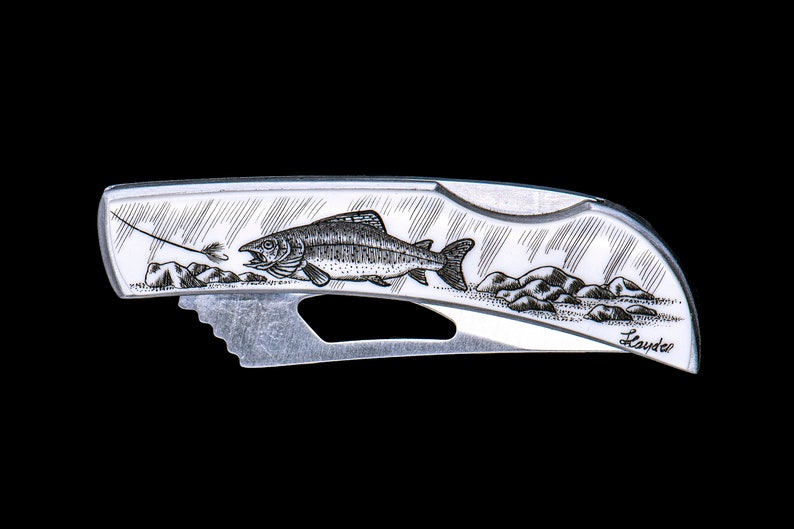 Peut inclure: Un couteau de poche argent&eacute; avec une gravure d&eacute;taill&eacute;e d'une truite sautant hors de l'eau. Le couteau a une petite lame et un manche avec le mot "Lauder" grav&eacute; dessus.