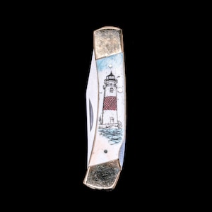 Könnte beinhalten: Ein Klappmesser mit dekorativem Griff, der einen Leuchtturm zeigt. Der Griff hat einen weißen Hintergrund mit einem rot-weißen Leuchtturm, blauem Himmel und goldfarbenen Metallakzenten. Die Klinge ist silberfarben.