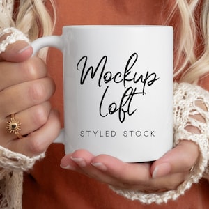 May include: A white ceramic mug with a black cursive inscription that reads "Mockup Loft" and the text "Styled Stock" below it. The mug is being held by a person wearing a white crocheted cardigan.