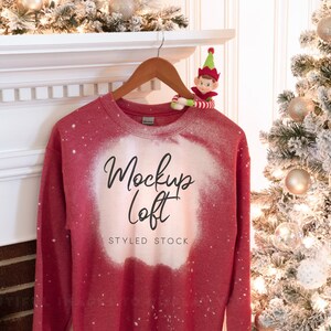 May include: Red bleached sweatshirt with the text "Mockup Loft" and "Styled Stock" on the front. The sweatshirt is hanging on a wooden hanger against a white fireplace mantel.
