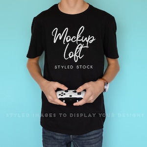 Peut inclure: Un t-shirt noir avec le texte "Mockup Loft" en écriture blanche. Une personne tient le t-shirt et une manette de jeu vidéo blanche et noire. Le fond est bleu clair uni.