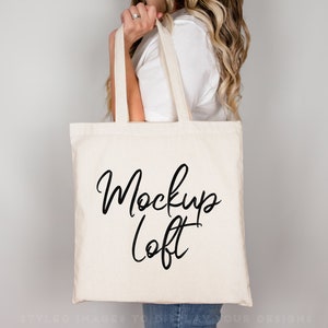 Puede incluir: Un bolso tote de color crema con las palabras "Mockup Loft" en escritura negra. El bolso tiene asas largas y lo sostiene una persona. El bolso est&aacute; hecho de una tela resistente.