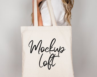 Puede incluir: Un bolso tote de color crema con las palabras "Mockup Loft" en escritura negra. El bolso tiene asas largas y lo sostiene una persona. El bolso está hecho de una tela resistente.