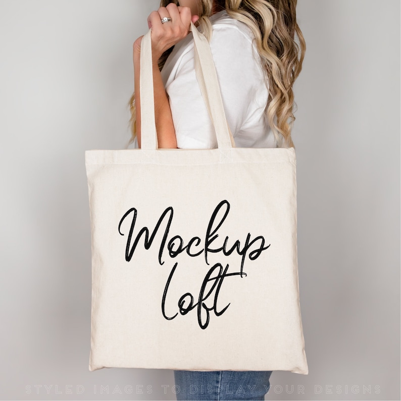 Puede incluir: Un bolso tote de color crema con las palabras "Mockup Loft" en escritura negra. El bolso tiene asas largas y lo sostiene una persona. El bolso est&aacute; hecho de una tela resistente.