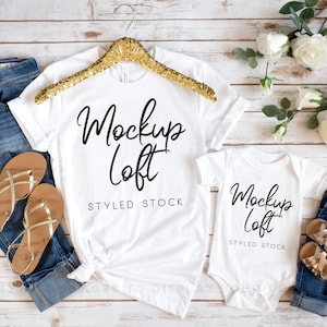 May include: Two white t-shirts with the text "Mockup Loft" and "Styled Stock" on a white wood background. The shirts are styled with denim jeans and sandals.