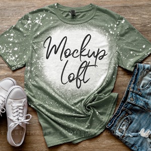 Può includere: Una maglietta verde con schizzi di candeggina bianca e il testo "Mockup Loft" in nero. La maglietta è stesa su una superficie di legno con un paio di scarpe da ginnastica bianche e un paio di pantaloncini di jeans blu.