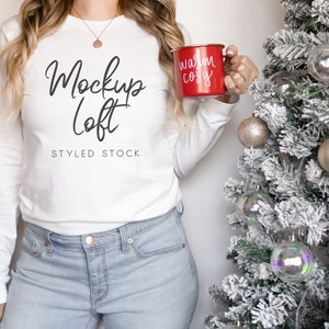 May include: A white long-sleeved shirt with the text "Mockup Loft" and "Styled Stock" printed on the front. The person is holding a red mug with the text "Warm Cozy" printed on it.