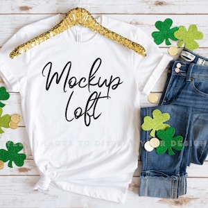 May include: White t-shirt with the text "Mockup Loft" in black script font. The shirt is on a wooden surface with a gold glitter hanger, green shamrock decorations, and gold coins.