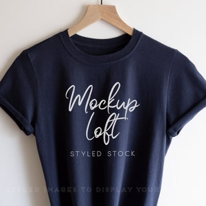 May include: A navy blue t-shirt with rolled up sleeves hanging on a wooden hanger. The shirt has the text "Mockup Loft" and "Styled Stock" printed on the front in white.
