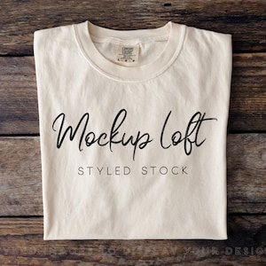 Puede incluir: Una maqueta de camiseta lisa de color blanco roto sobre un fondo de madera. El texto "Mockup Loft" y "Styled Stock" está impreso en la camiseta.