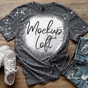 May include: A dark gray t-shirt with white bleach spots and the text "Mockup Loft" in a script font. The shirt is paired with white sneakers and denim shorts on a wooden surface.