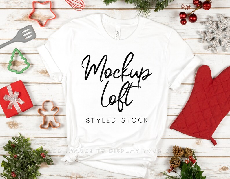 Pu&ograve; includere: Mockup di una t-shirt bianca con il testo "Mockup Loft" e "Styled Stock" su uno sfondo di legno bianco con decorazioni natalizie.