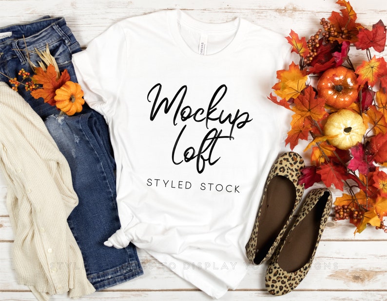 May include: White t-shirt mockup with the text "Mockup Loft" and "Styled Stock" on a white wooden background. The t-shirt is styled with blue jeans, a cream cardigan, and leopard print flats. Fall leaves and pumpkins are in the background.