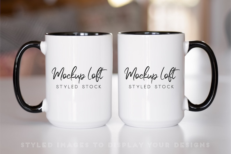 Może przedstawiać: Dwa białe ceramiczne kubki z czarnymi uchwytami i obw&oacute;dkami. Kubki mają napis "Mockup Loft STYLED STOCK" w odręcznej czcionce. Kubki są prezentowane na białej powierzchni.