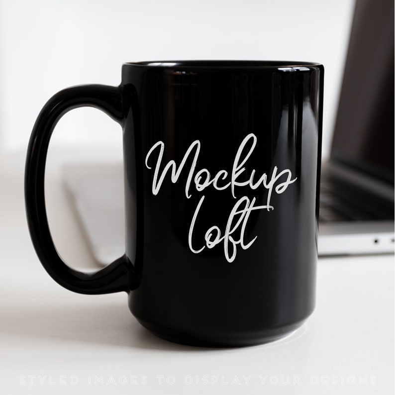 Pu&ograve; includere: Una tazza in ceramica nera con una scritta bianca che recita "Mockup Loft". La tazza &egrave; appoggiata su una superficie bianca accanto a un laptop.