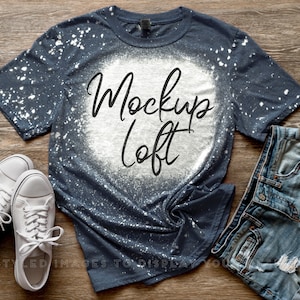Könnte beinhalten: Ein blaues T-Shirt mit weißen Bleichmittelspritzern und dem Text "Mockup Loft" in Schwarz. Das Shirt ist zerknittert und liegt auf einer Holzoberfläche mit einem Paar weißen Turnschuhen und einer blauen Jeans-Shorts.