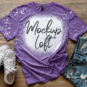 May include: Purple t-shirt with white bleach splatters and the text "Mockup Loft" in black. The shirt is styled with denim shorts and white sneakers.