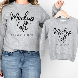 May include: Two grey sweatshirts with the text "Mockup Loft" and "STYLED STOCK" on them. The larger sweatshirt is being worn by a person, while the smaller sweatshirt is hanging on a wooden hanger.