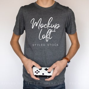 May include: A person wearing a gray t-shirt with the text "Mockup Loft STYLED STOCK" printed on the front. The person is holding a white video game controller.