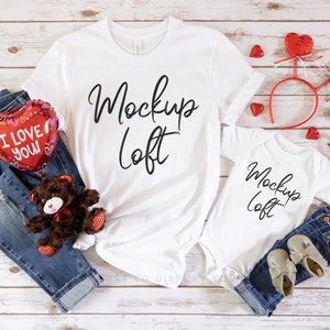May include: White t-shirts with the text "Mockup Loft" on them. The shirts are laid out on a white wooden surface with red heart-shaped decorations, a teddy bear, and jeans.