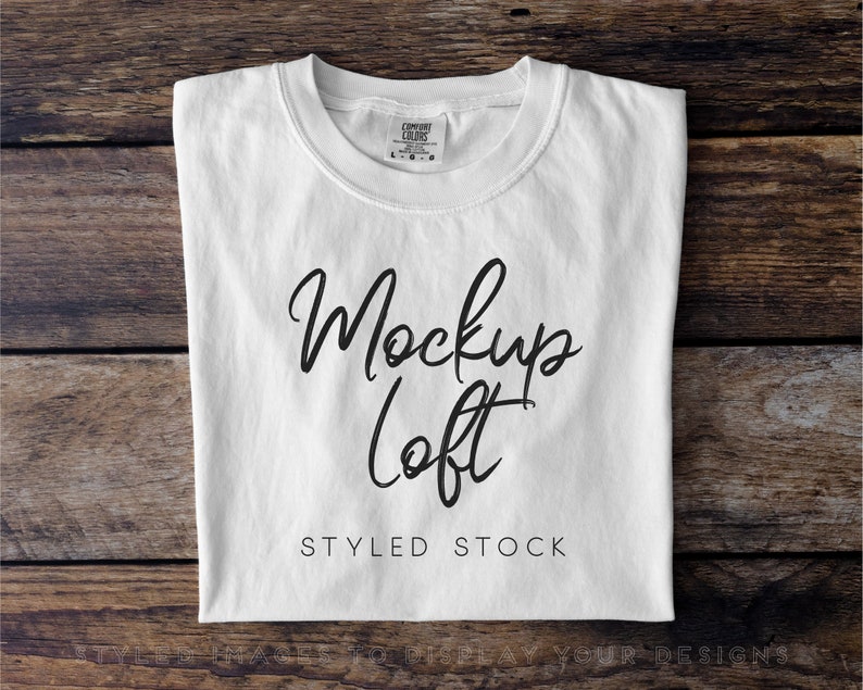 Op de afbeelding: Witte t-shirt mockup op een houten achtergrond. Het t-shirt is gevouwen en heeft de tekst "Mockup Loft" en "Styled Stock" erop gedrukt.