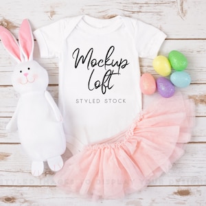 May include: White baby onesie with the text "Mockup Loft" and "Styled Stock" on a white wooden background. A pink tutu, a white stuffed bunny, and pastel coloured Easter eggs are also in the image.