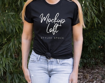 May include: A black t-shirt with the text "Mockup Loft" and "Styled Stock" in white lettering. The shirt is being worn by a person with long blonde hair and is tied at the waist.
