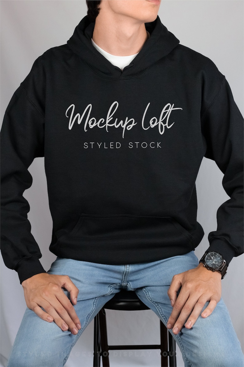 May include: A black hoodie with a front pocket and a white script that reads "Mockup loft STYLED STOCK". The model is wearing light blue jeans and a watch with a black band and face.