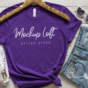 May include: Purple t-shirt mockup with a gold glitter hanger, white converse shoes, denim shorts, and white heart-shaped sunglasses. The t-shirt has the text "Mockup Loft Styled Stock" printed on it.