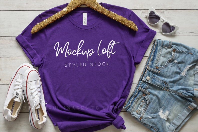 May include: Purple t-shirt mockup with a gold glitter hanger, white converse shoes, denim shorts, and white heart-shaped sunglasses. The t-shirt has the text "Mockup Loft Styled Stock" printed on it.