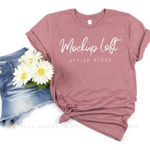May include: A dusty rose t-shirt with the text "Mockup Loft STYLED STOCK" in white script. The shirt is styled with denim shorts and white flowers. The shirt is knotted at the bottom.