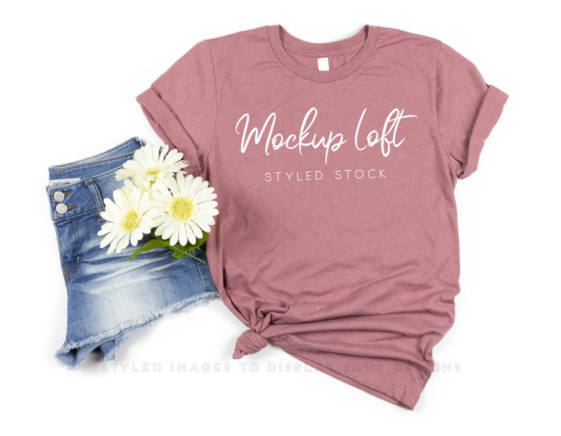 May include: A dusty rose t-shirt with the text "Mockup Loft STYLED STOCK" in white script. The shirt is styled with denim shorts and white flowers. The shirt is knotted at the bottom.