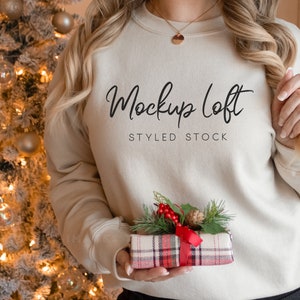 May include: A woman wearing a light beige sweatshirt with the text "Mockup Loft Styled Stock" on the front. She is holding a small gift box wrapped in red and black plaid paper with a red ribbon.