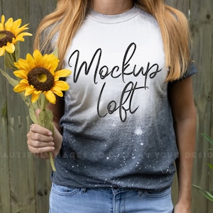 May include: A woman wearing a gray and white tie-dye t-shirt with the text "Mockup Loft" printed on the front. She is holding a bouquet of yellow sunflowers in front of a wooden fence.