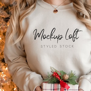 K&ouml;nnte beinhalten: Ein wei&szlig;er Pullover mit dem Aufdruck "Mockup Loft STYLED STOCK" auf der Vorderseite. Der Pullover wird von einer Person getragen, die ein verpacktes Geschenk mit einer roten Schleife h&auml;lt.
