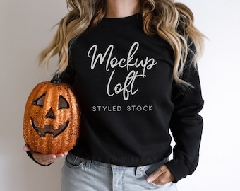 May include: A woman wearing a black sweatshirt with the text "Mockup Loft Styled Stock" in white. She is holding a glittery orange pumpkin with a jack-o'-lantern face.