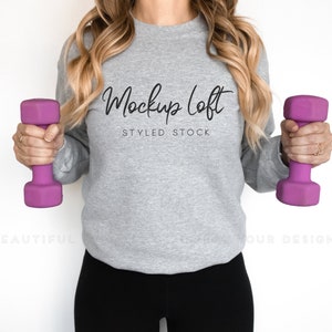 May include: A woman wearing a gray sweatshirt with the text "Mockup Loft Styled Stock" and black leggings, holding a purple dumbbell in each hand.